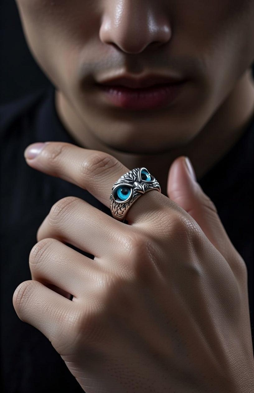 A person with softly parted lips wears a silver ring shaped like an owl with blue gemstone eyes, prominently displayed on their index finger against a dark background.